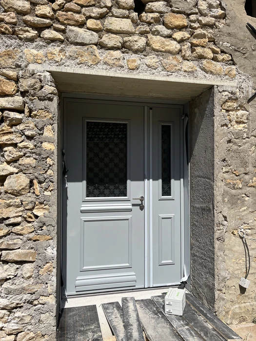 Entrée d'un bâtiment ancien en pierre de taille dorée avec porte d'accès moderne bicolore blanc et gris, comportant un vitrage supérieur à motif géométrique noir et une partie inférieure opaque