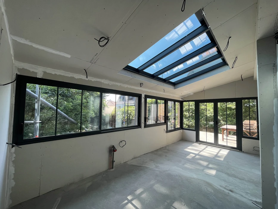 Intérieur en cours de finition d'une véranda ou extension résidentielle avec multiples baies vitrées à cadres noirs offrant une vue sur jardin paysagé