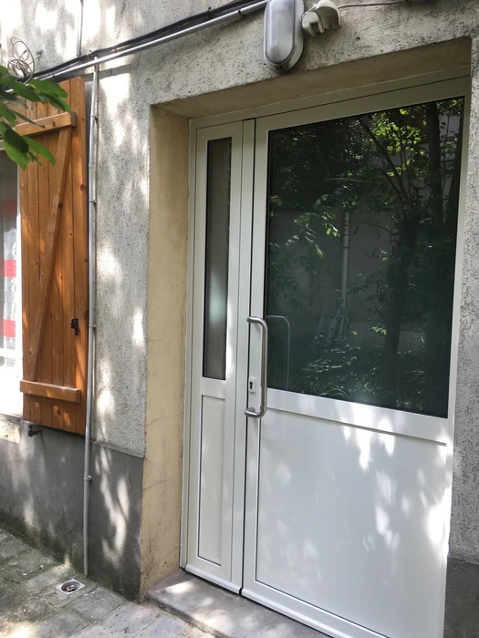 Entrée d'immeuble avec porte-fenêtre vitrée blanche et bois, bardage partiel blanc inférieur, encadrement beige clair, mur gris ciment, porte bois teinté à gauche, interphone gris en applique, poignées chromées, vitrage teinté réfléchissant, visible végétation extérieure