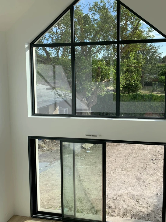 Menuiserie en tenue de travail grise relevant avec un mètre ruban la largeur d'une ouverture murale dans un salon rénové, avant la fabrication d'une verrière en acier noir sur mesure, vue de face, lumière naturelle.