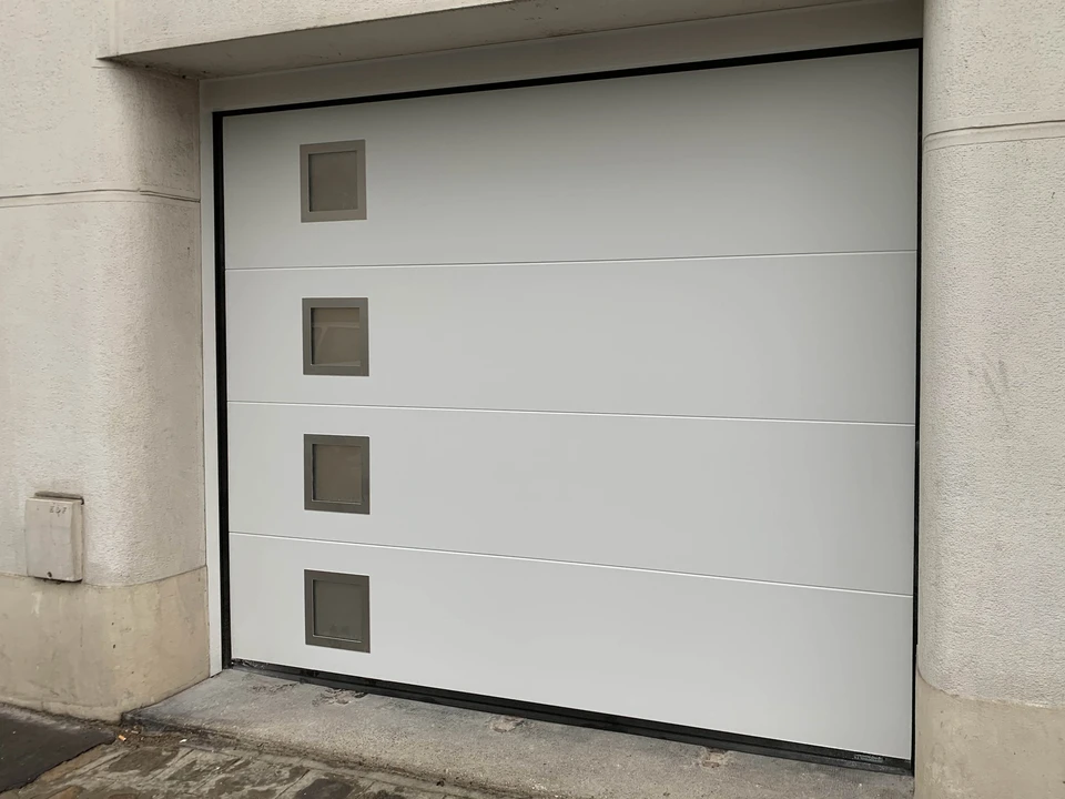 Artisan menuisier en tenue de travail relevant avec un mètre ruban la largeur de l'ouverture d'un garage de maison individuelle, carnet de notes à la main, vu de l'extérieur, lumière du jour.