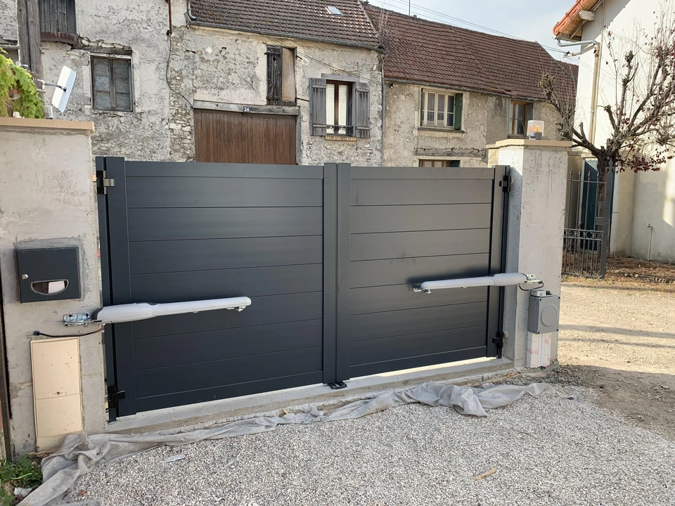Artisan menuisier en tenue de travail, un mètre ruban tendu devant un portail aluminium anthracite entre deux piliers maçonnés, tablette de notes à la main, lors d'une visite technique sur place chez un particulier.
