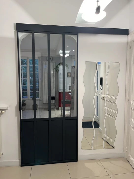 Vue ext""erieure d'une porte-fen""etre cintr""ee en PVC blanc ""a petits carreaux et imposte ovale, install""ee dans une fa""cade en pierre de taille, terrasse en bois composite, jardini""eres de fleurs rouges, lumi""ere naturelle du jour.